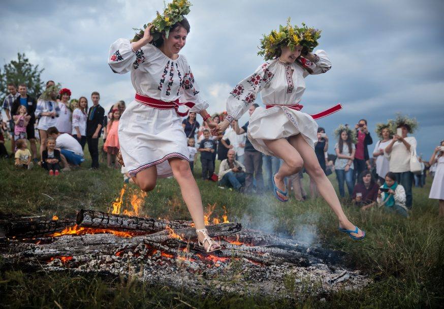 Due donne vestite di bianco con una cinta rossa e con una ghirlanda di fiori gialli e verdi sul capo attraversano una brace