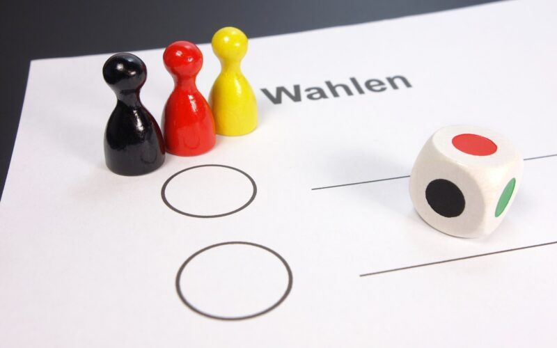 foglio per le elezioni con sopra un dado e tre pedoni colorati come la bandiera tedesca